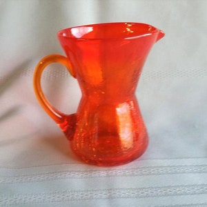 May include: A vibrant orange glass pitcher with a handle and a pouring spout. The pitcher has a textured surface and a rounded base. The handle is a lighter shade of orange. The pitcher is likely used for serving beverages.