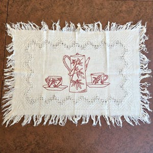 May include: A rectangular off-white placemat featuring a fringed edge and a decorative openwork border. The center displays a red embroidered design of a coffee pot and two matching cups on saucers, all decorated with a bamboo leaf pattern.