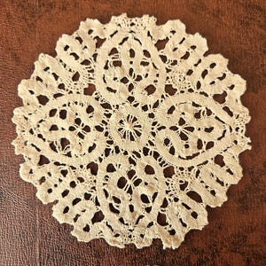 May include: A cream-colored, circular doily with an intricate lace design. The doily features a central floral motif surrounded by repeating patterns and a scalloped edge. The delicate openwork creates a textured appearance.