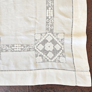 May include: Close-up of a white linen tablecloth with intricate lace detailing. The tablecloth features a geometric lace pattern in the corners and along the edges, with a delicate stitched border. The fabric has a subtle texture.