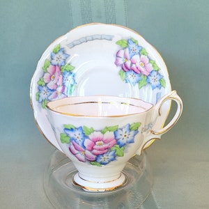 May include: A white teacup and saucer set with a floral design in pink and blue. The teacup and saucer have gold trim. The teacup is on a clear glass stand. The background is a solid light green color.