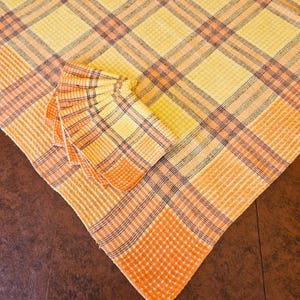 May include: A yellow and orange plaid tablecloth with matching napkins. The tablecloth and napkins have a classic plaid pattern in shades of yellow, orange, and a hint of purple. The napkins are neatly folded and stacked on the tablecloth.