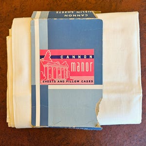 May include: A vintage package of Cannon Manor sheets and pillowcases. The packaging is blue and white with a pink label featuring an illustration of a building. The sheets are a light cream color, and the package is slightly torn.