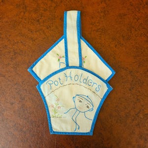May include: Cream-colored pot holders with blue trim and a handle. The pot holders have embroidered details, including the words "Pot Holders" and a whimsical figure. Pink floral accents are also present.