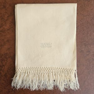 May include: A rectangular cream-colored textile featuring a delicate, tonal embroidered monogram in the center. The bottom edge is finished with a decorative, hand-knotted macramé fringe detail that extends downward in loose, fine threads.
