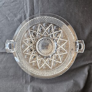 May include: Clear glass serving tray with a starburst design in the center. The tray has a circular shape with two rectangular handles on opposite sides. The rim features a textured pattern.