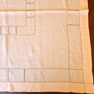 May include: Cream-colored linen tablecloth with a geometric design. The edges and the interior design elements are outlined with a dark brown, dotted stitch. The tablecloth is rectangular and has a simple, elegant aesthetic.