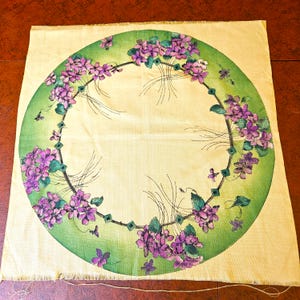 May include: A cream-coloured square fabric featuring a large, circular floral design. The design has a green border with a ring of purple flowers and green leaves. Small purple butterflies are scattered around the floral ring.