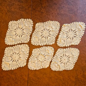 May include: Six cream-colored, crocheted doilies arranged on a brown surface. Each doily has a square shape with a scalloped edge and a detailed, openwork floral pattern. The doilies are handmade and have a vintage aesthetic.