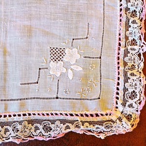 May include: Close-up of a white handkerchief with delicate floral embroidery and a decorative lace border. The handkerchief features a pink trim and intricate stitching details, showcasing a vintage aesthetic.