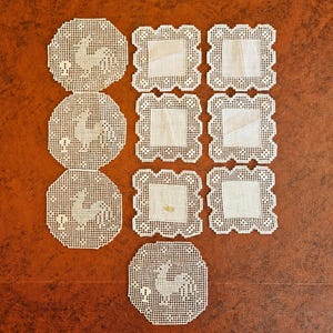 May include: A collection of handmade, off-white coasters and doilies. The coasters are round with a rooster design, while the doilies are square with a scalloped edge. The items are arranged on a brown surface.