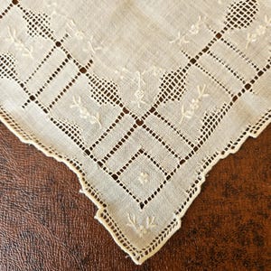 May include: Close-up of a vintage white handkerchief with intricate embroidered floral and geometric patterns. The handkerchief features delicate openwork and scalloped edges, showcasing a classic, elegant design. The fabric appears to be linen or a similar fine weave.