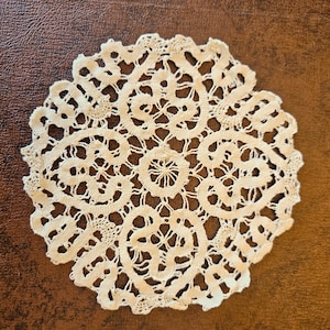 May include: Cream-colored, circular doily with an intricate lace pattern. The design features a central floral motif surrounded by swirling, openwork details. The edges are scalloped, adding a delicate touch to the handmade textile.