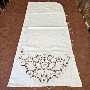 May include: Cream-colored, rectangular textile with an intricate, openwork floral design along the bottom edge. The fabric appears to be linen or a similar material, with a slightly textured surface. The piece is laid out on a brown surface.