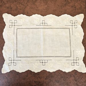May include: A rectangular, off-white linen doily with a scalloped edge. The doily features embroidered geometric and floral patterns in a darker thread, including squares, triangles, and stylized flowers. The central area is plain, framed by a dotted border.
