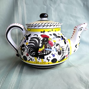 May include: A hand-painted ceramic teapot with a rooster design. The teapot features a black rooster with a red comb and yellow accents on a white background. The handle and spout are white with black and red details. The lid has a black knob.