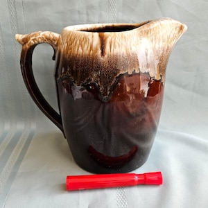 May include: A ceramic pitcher with a glossy dark brown base and a cream-colored drip glaze at the rim. The pitcher features a curved handle and a pouring spout. A red cylindrical object is positioned in front for scale, measuring 3.5 inches long.