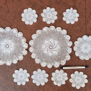May include: A collection of handmade, white crochet doilies and coasters arranged on a brown surface. The doilies vary in size, with a large central piece and smaller, star-shaped coasters. The intricate lace patterns are visible.