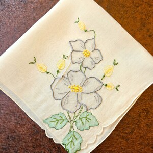 May include: Cream-colored handkerchief with embroidered floral design. Features two large white flowers with yellow centers and gray outlines, along with green leaves and yellow buds. Scalloped edge adds a delicate touch. A vintage-inspired accessory.