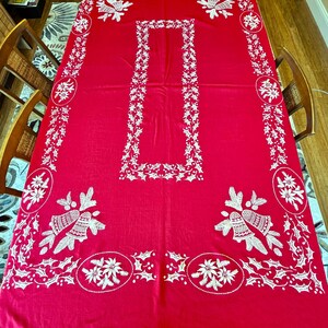 May include: A red rectangular tablecloth with white embroidered floral and bell designs. The tablecloth features a central rectangular border and decorative patterns along the edges, creating a festive holiday theme. The tablecloth is made of a soft fabric.