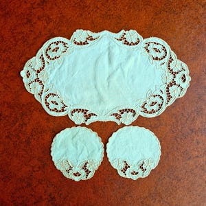 May include: A light blue, oval-shaped doily with floral cutouts and two matching round coasters. The edges of the doily and coasters feature delicate floral embroidery. The set is made of a soft, possibly linen, material.
