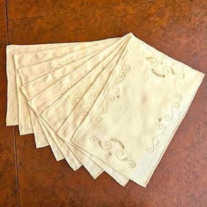 May include: A set of six cream-colored rectangular placemats. Each placemat features an embroidered design of swirling lines and dots in a matching cream color. The placemats are arranged in a fan shape on a brown surface.
