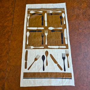 May include: A rectangular kitchen towel featuring a brown and white color scheme with a repeating pattern of black and white cutlery silhouettes including forks, knives, and spoons arranged in a grid and scattered across the fabric surface.