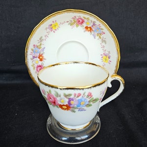 May include: A white teacup and saucer set with a floral design. The cup and saucer have gold trim and a colorful floral pattern of pink, blue, yellow, and orange flowers. The teacup is on a clear stand.