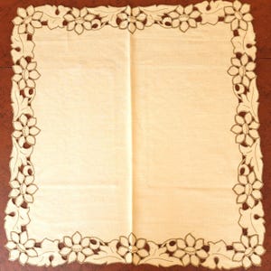 May include: A square cream-colored textile mat featuring a decorative border of cutwork embroidery. The border consists of repeating floral and leaf motifs with intricate stitching along the edges, framing a plain, solid-colored central panel.