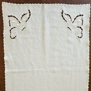 May include: Cream-colored rectangular linen doily with two butterfly cutouts. The butterflies are near the top corners and have detailed wing designs. The edges are finished with a delicate, scalloped trim. The doily is likely handmade.