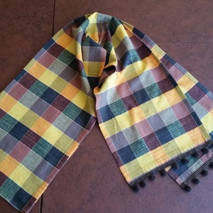 May include: A multi-coloured plaid scarf featuring a repeating pattern of squares in yellow, green, brown, and blue. The scarf has a black pom-pom trim along the edges. The scarf is made of a soft fabric.