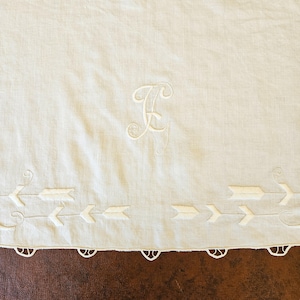 May include: Cream-colored linen textile with embroidered details. Features a large, stylized letter "F" in the upper center and a row of embroidered arrows along the bottom edge. The bottom edge has a scalloped trim.