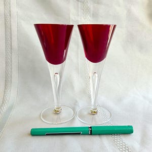 May include: Two elegant wine glasses with ruby red bowls and clear stems and bases. The glasses are next to a green pen. The glasses are likely for serving wine or other beverages.