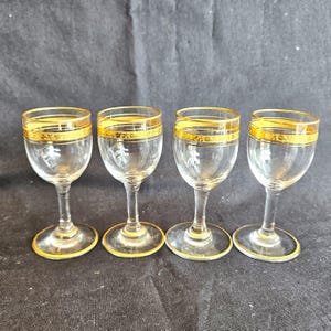 May include: A set of four clear glass stemmed glasses, each featuring a gold rim and a decorative gold band around the upper bowl. The glasses have a slender stem and a circular base with a thin gold trim along the edge of the foot.