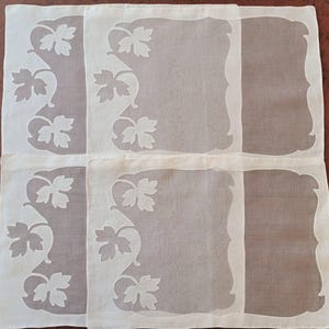 May include: A set of four square placemats made of sheer off-white fabric. Each placemat features a decorative border with a scalloped edge and a repeating pattern of white vine leaves on the left side, creating a semi-transparent effect.