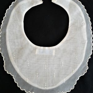 May include: A white, sheer, semi-circular bib featuring a scalloped edge with delicate white embroidery. The fabric is lightweight and translucent, decorated with small, evenly spaced white bead accents arranged in vertical rows across the front.