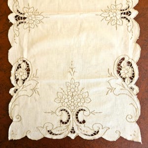 May include: Cream-colored embroidered table runner with a scalloped edge. The runner features floral and scrollwork designs in a matching thread color. The embroidery includes cut-out details, adding a delicate touch to the textile.