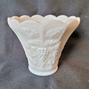 Peut inclure: Un vase blanc de style vintage avec une forme évasée et un bord festonné. Le vase présente des motifs de raisins et de feuilles en relief. La base est ronde et le vase est en verre blanc laiteux. Le vase mesure environ 15 cm de haut.