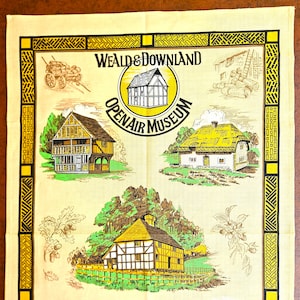 May include: A light yellow tea towel featuring illustrations of historic buildings and the text "Weald & Downland Open Air Museum." The design includes a decorative border in black and yellow, with drawings of a cart, trees, and foliage.
