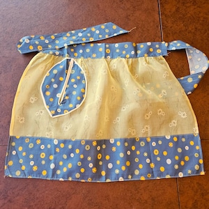 May include: A half apron featuring a light yellow fabric with a white daisy print, accented by a blue waistband and bottom border patterned with yellow and white polka dots. A single leaf-shaped pocket with matching polka dots is attached.