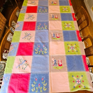 May include: A patchwork tablecloth with a variety of colors including pink, blue, green, and gray. The squares are stitched together with white thread. Embroidered birds, floral designs, and figures are featured on the tablecloth.