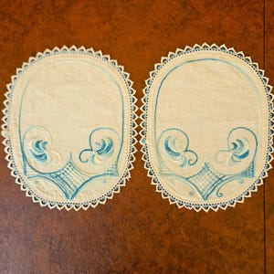 May include: Two oval-shaped, cream-colored embroidered doilies with blue floral designs and scalloped edges. The embroidery features swirling patterns and geometric shapes, creating a vintage aesthetic. The doilies are placed side by side.
