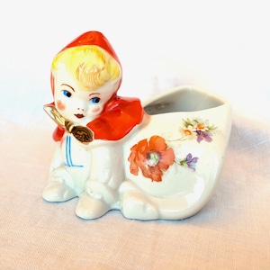 May include: A vintage ceramic figurine of a child in a red hooded cape, holding a paintbrush, next to a white clog planter. The clog is decorated with a red poppy and other floral designs. The figurine is painted with bright colors.
