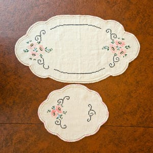 May include: Two off-white embroidered placemats with scalloped edges. The larger mat has a floral design with pink and green accents, and a black stitched border. The smaller mat has a similar design. The mats are on a brown surface.