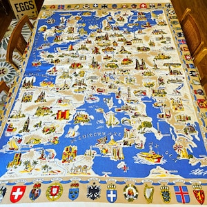 May include: A vintage tablecloth featuring a detailed map of Europe with illustrations of landmarks and cities. The map is set against a blue and tan background, with a border of flags and crests. The tablecloth is spread out on a wooden table.