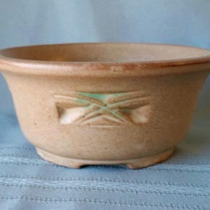 Peut inclure: Un bol en céramique de couleur beige, doté d'un bord évasé et d'une base surélevée. L'extérieur présente un motif géométrique en relief au centre, avec des touches de glaçure verte dans les lignes creuses de la surface en argile mate.