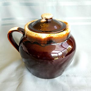 May include: A round ceramic sugar bowl with a glossy dark brown glaze and a cream-coloured drip pattern along the rim and lid. The bowl features a single curved handle on the left side and a matching lid with a small knob handle on top.
