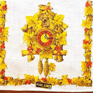 May include: A white linen tea towel featuring a detailed illustration of a cuckoo clock. The clock is brown and gold, surrounded by yellow leaves and red berries. The clock face has Roman numerals. The border is decorated with leaves and berries.