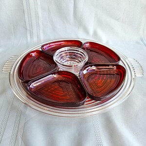 May include: A clear glass serving tray with two side handles, containing four deep red, triangular glass dishes arranged around a central, clear glass bowl. The red dishes feature a textured, geometric pattern on their undersides.
