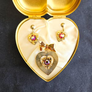 May include: A vintage gold-toned jewelry set, including heart-shaped earrings and a pendant necklace, presented in a heart-shaped box. The earrings and pendant feature a floral design with iridescent crystals. The box is lined with cream-colored satin.